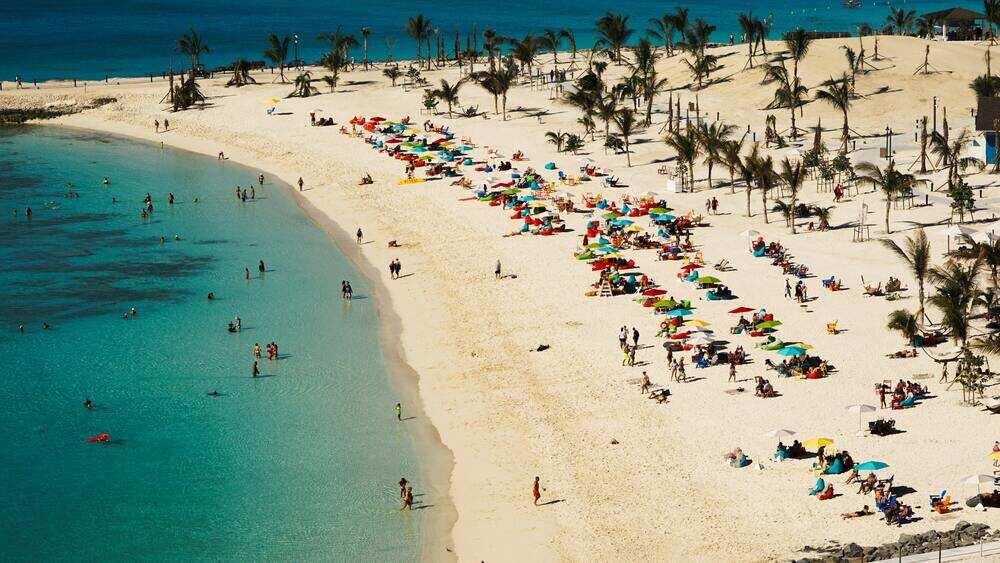 People swimming at the beach and some are standing, sitting or laying down at the white sand with green trees 