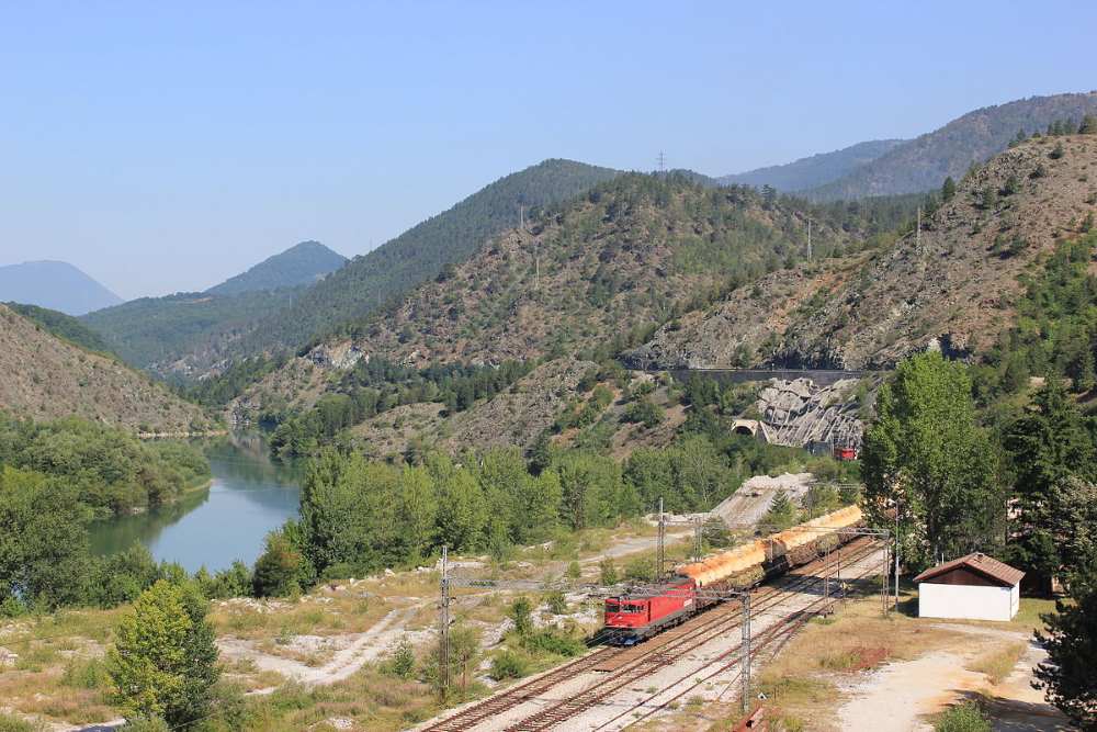 aerial view of a red train passing by  beside is body of water and lushy mountains