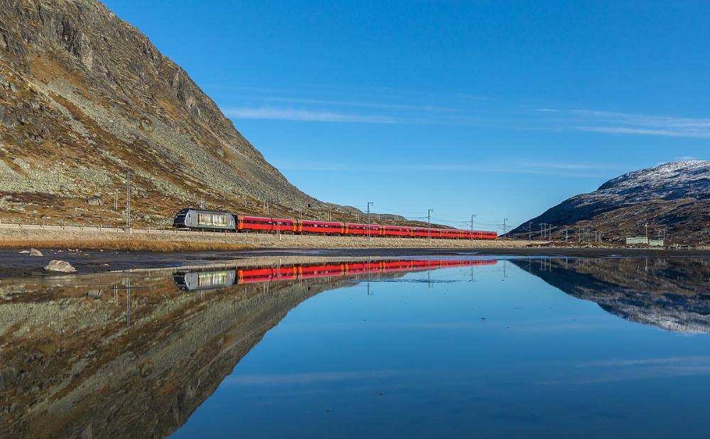 red train passing by near mountain and body of water - most beautiful train rides in the world