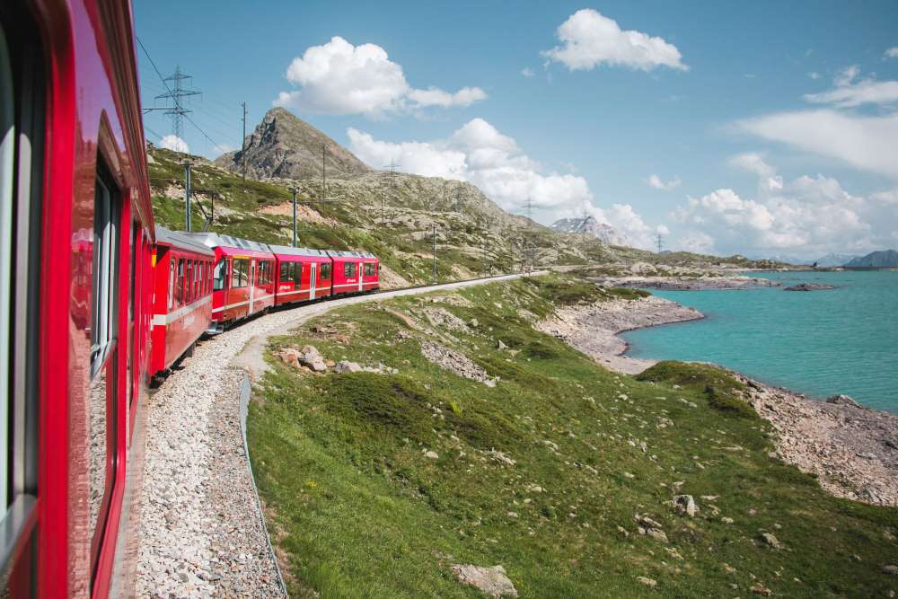 red train passing by near body of water - most beautiful train rides in the world