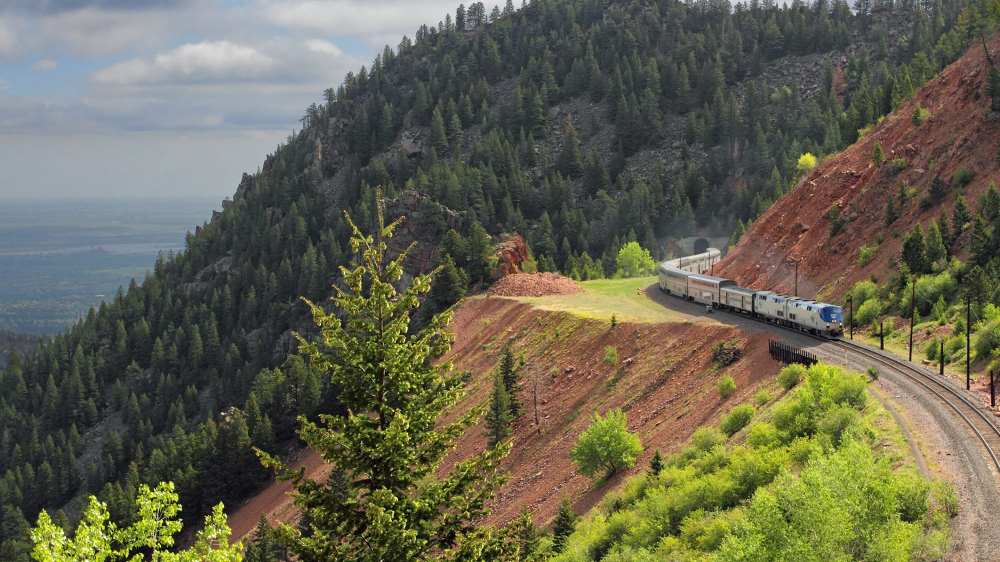train passing by narrow trail surrounded by trees and mountains - most beautiful train rides in the world