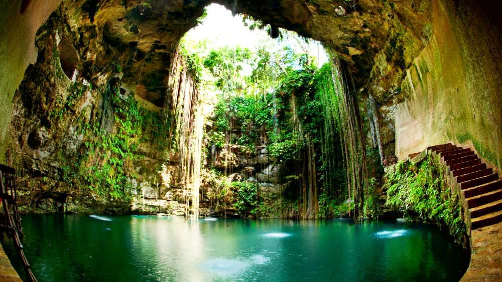 This open-air cenote is a large sinkhole filled with crystal-clear, turquoise water, surrounded by lush vegetation and towering limestone walls.