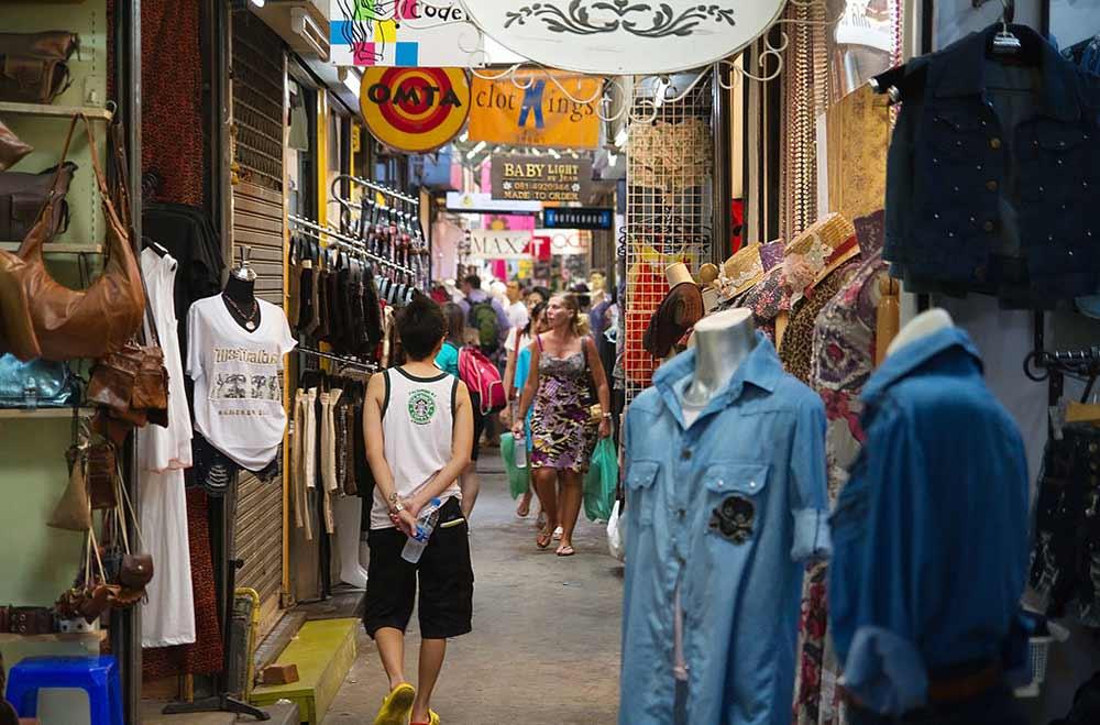 people walking around a street market with variety of clothes and bags for sale