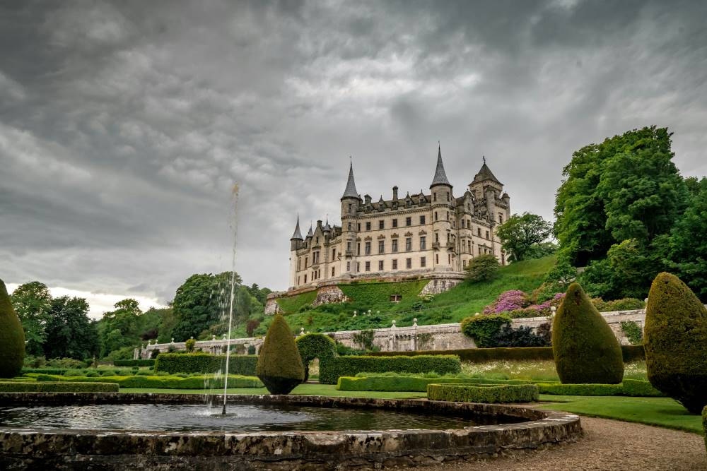 white concrete castle on top surrounded by plants and trees with fountain in front