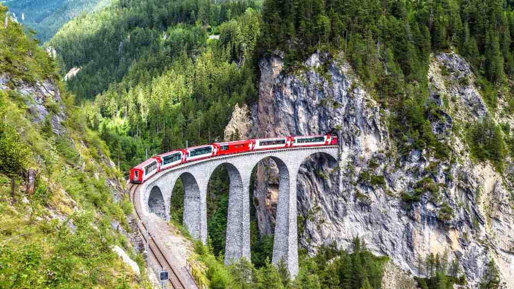 aerial view of red train passing by tunnel surrounded by lushy green mountain - most beautiful train rides in the world
