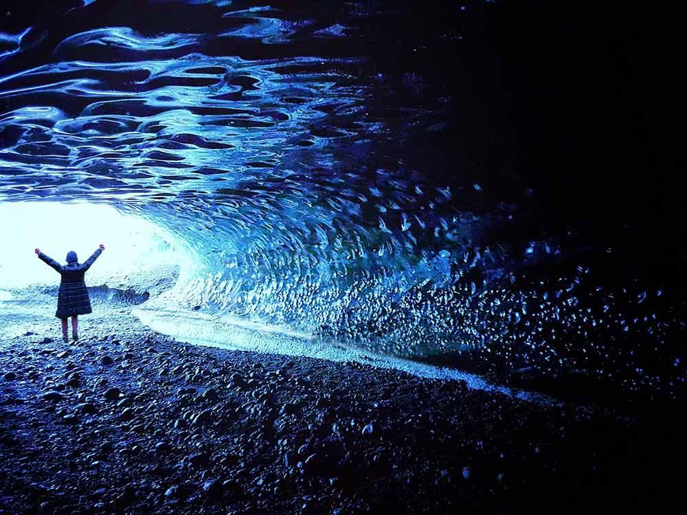 back of a woman in jacket raising both arms in the air inside the glacier man made tunnel