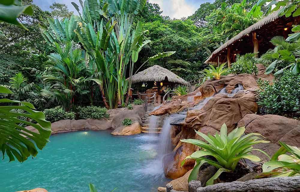 Calming view of a hot springs surrounded by green leafy trees and a small nipa hut with brown stone on the side of the spring