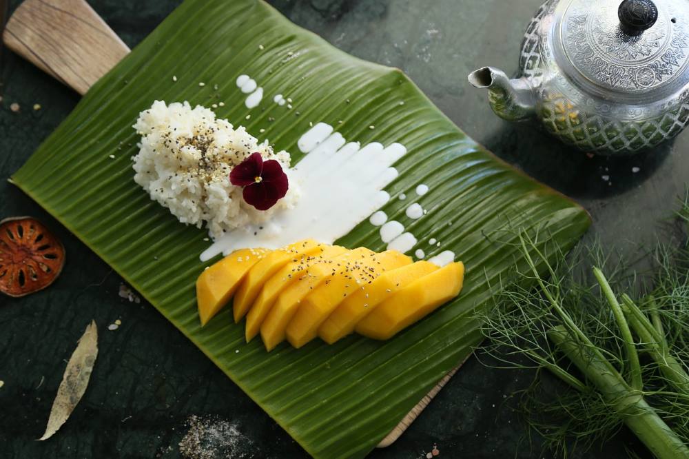 mango sticky rice on top of green leaf plate and beside is silver kettle