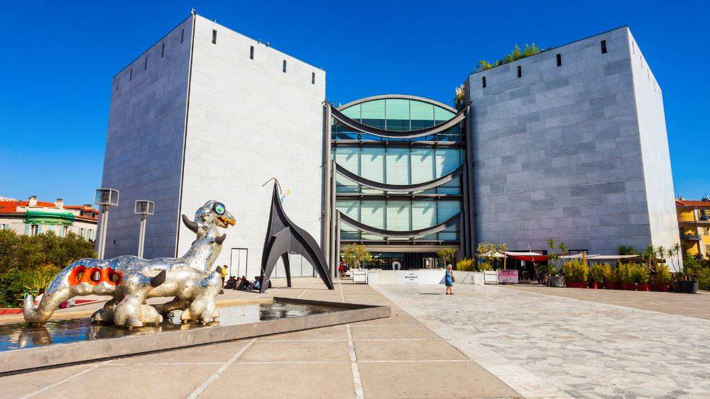 two gray building connected with one in the middle made of glass building and outside is a statue on top of a water