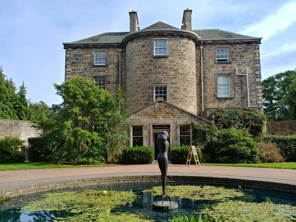 old structure building surrounded by tress and plants in front of a pond with statue of a body without head- Things to Do in Scotland