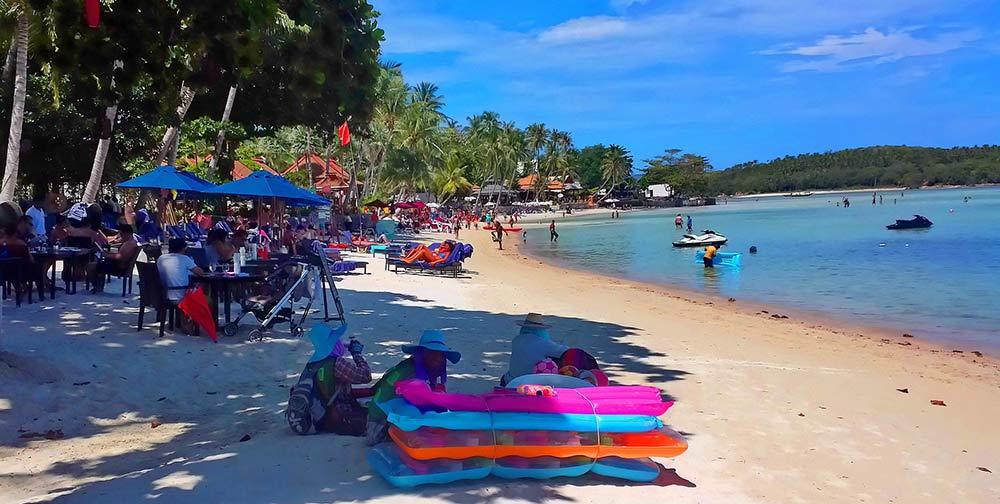 Crowded beach with people sitting at the table and chairs beside the beach