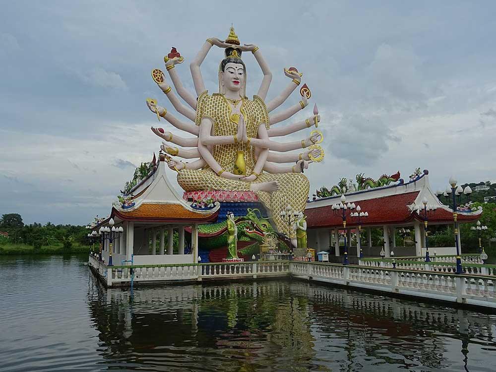 big buddha with a lot of arms on top of a little building floating in the beach side