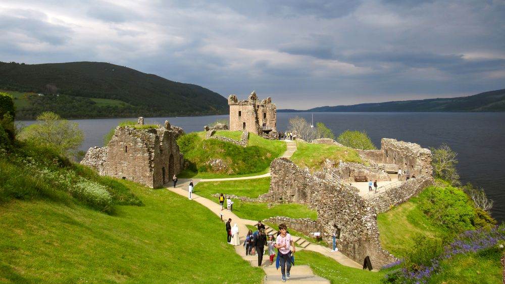 people walking in a green hill surrounded by ruin rock structure beside body of water- Things to Do in Scotland