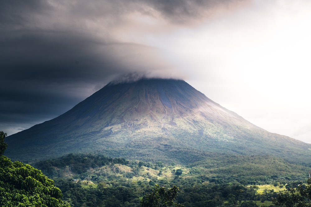 A perfect cone volcano with clouds on top and green valley below