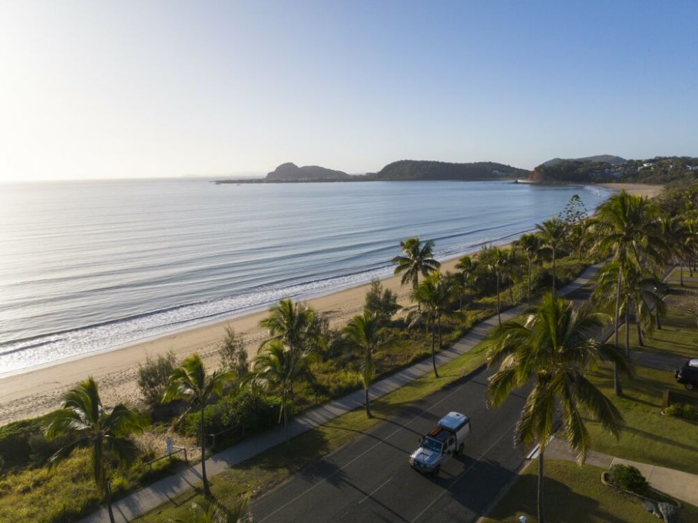 vehicle strolling in an empty road surrounded by palm tree with beach on the side - Best Places To Visit In Queensland
